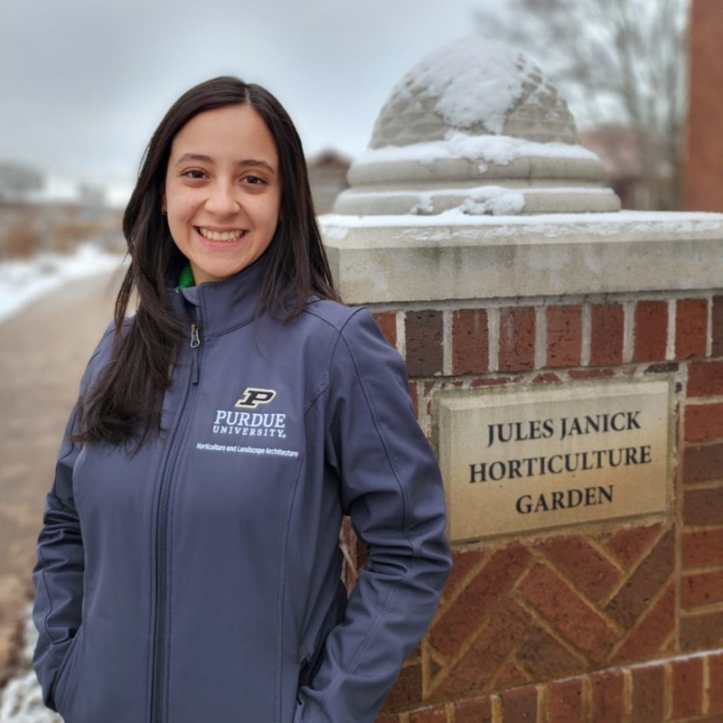 Lian Durón Alvarado standing in front of the Jules Janice Horticulture Garden, wearing a Purdue University jacket.